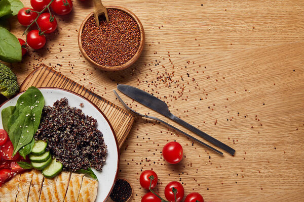 top view of cooked quinoa with grilled chicken breast and vegetables on wooden table with cutlery