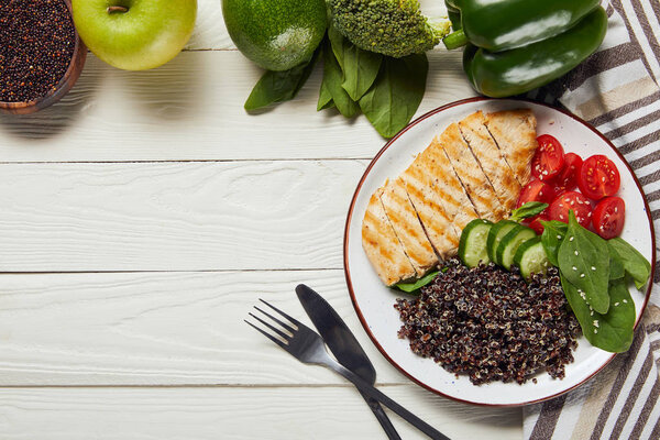 top view of cooked quinoa with grilled chicken breast and vegetables on white wooden table with cutlery and copy space