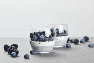 selective focus of tasty yogurt with chia seeds, blueberries and blackberries in glasses near scattered berries on marble surface isolated on grey clipart