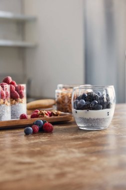 selective focus of glasses with yogurt, oat flakes and berries on wooden tray on table in kitchen clipart
