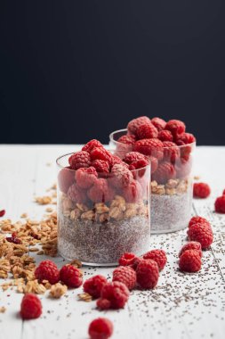 selective focus of glasses with yogurt, oat flakes and raspberries near scattered chia seeds, oat flakes and berries on white wooden table isolated on black clipart