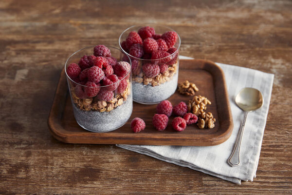 tasty yogurt with chia seeds, oat flakes, nuts and raspberries on wooden tray on napkin with teaspoon on table