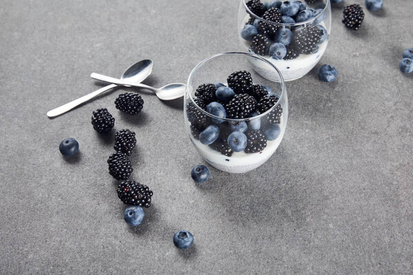 tasty yogurt with chia seeds, blueberries and blackberries in glasses near teaspoons and scattered berries on marble surface 
