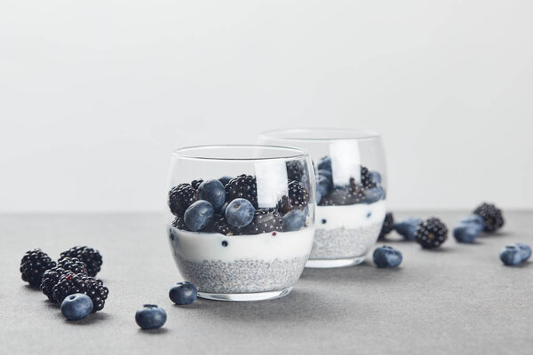 selective focus of tasty yogurt with chia seeds, blueberries and blackberries in glasses near scattered berries on marble surface isolated on grey