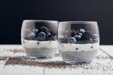 selective focus of yogurt with chia seeds, blueberries, blackberries near scattered seeds on white wooden isolated on black clipart
