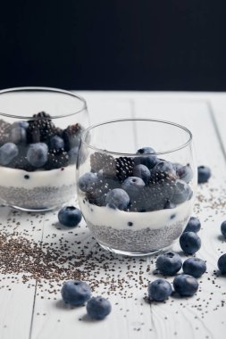 selective focus of tasty yogurt with chia seeds, blueberries, blackberries near scattered seeds and berries on white wooden isolated on black clipart