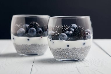 selective focus of yogurt with chia seeds, blueberries, blackberries on white wooden isolated on black clipart