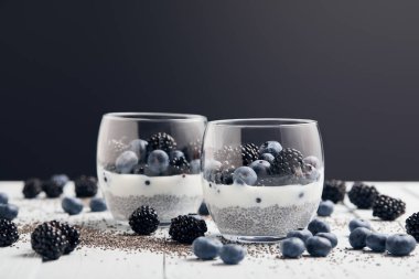 selective focus of yogurt with chia seeds and berries in glasses near scattered seeds, blueberries and blackberries on white table isolated on black clipart