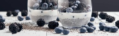 panoramic shot of yogurt with chia seeds and berries in glasses near scattered seeds, blueberries and blackberries on white table isolated on black clipart