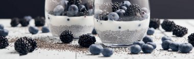 panoramic shot of yogurt with chia seeds and berries in glasses near scattered seeds, blueberries and blackberries on white table isolated on black clipart