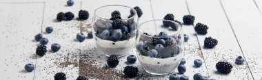 panoramic shot of tasty yogurt with chia seeds, blueberries, blackberries near scattered seeds and berries on white wooden isolated on black clipart