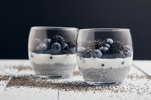 selective focus of yogurt with chia seeds, blueberries, blackberries near scattered seeds on white wooden isolated on black