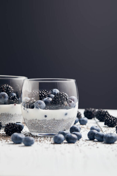 selective focus of tasty yogurt with berries near scattered chia seeds and berries on white wooden isolated on black 