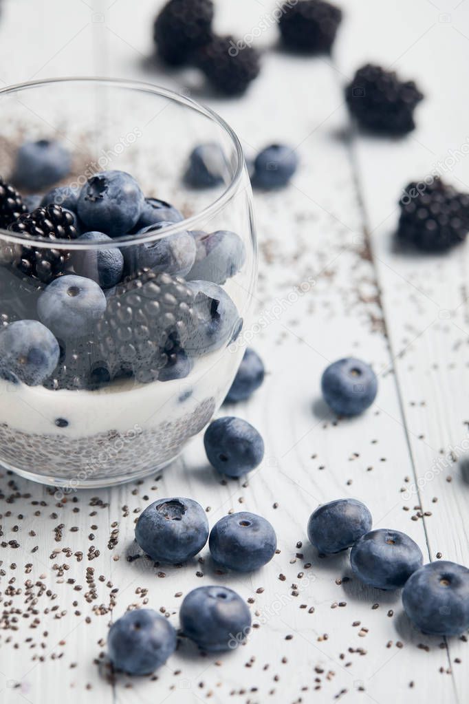 selective focus of blackberries, blueberries and yogurt in glass near scattered chia seeds and berries on white wooden table