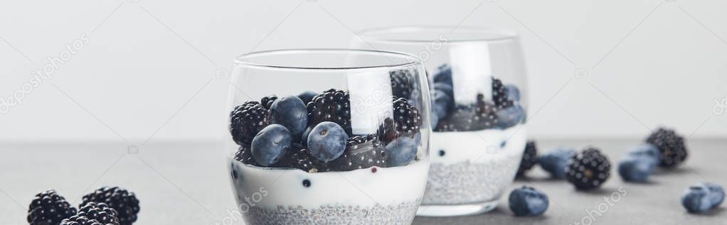 panoramic shot of tasty yogurt with chia seeds, blueberries and blackberries in glasses near scattered berries on marble surface isolated on grey