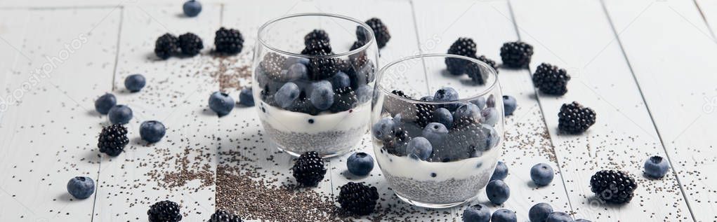 panoramic shot of tasty yogurt with chia seeds, blueberries, blackberries near scattered seeds and berries on white wooden isolated on black