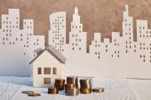 kovové mince a model domu na bílém dřevěném stole blízko hnědého texturované zdi, koncepce realit