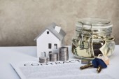 model domu na bílém stole se stříbrnými mincemi, klíči, zakázkou a pokladním políčky na šedé pozadí, koncept nemovitostí
