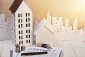 houses models on white wooden table with contract, coins and keys near white paper cut city, real estate concept
