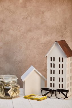 Beyaz ahşap masa üzerinde ev modelleri moneybox, hesap makinesi ve kahverengi dokulu duvar yakın gözlük, gayrimenkul kavramı
