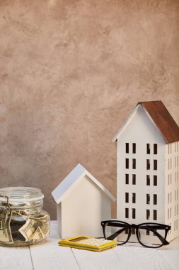 houses models on white wooden table with moneybox, calculator and glasses near brown textured wall, real estate concept clipart