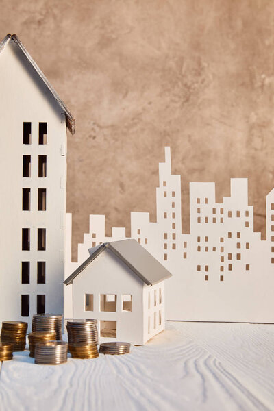 Монеты и дома модели на белом деревянном столе возле коричневой текстурированной стены и бумажного города, концепция недвижимости
