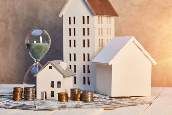 модели домов, монеты и наличные, песочные часы на белом деревянном столе с солнечным светом, концепция недвижимости
