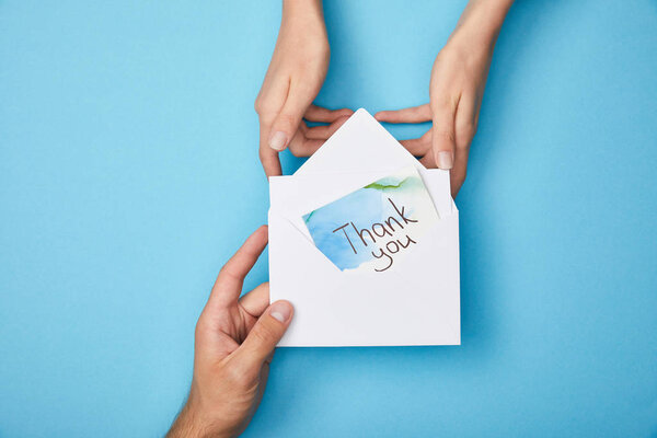 cropped view of man giving greeting card with thank you lettering in envelope to woman on blue background
