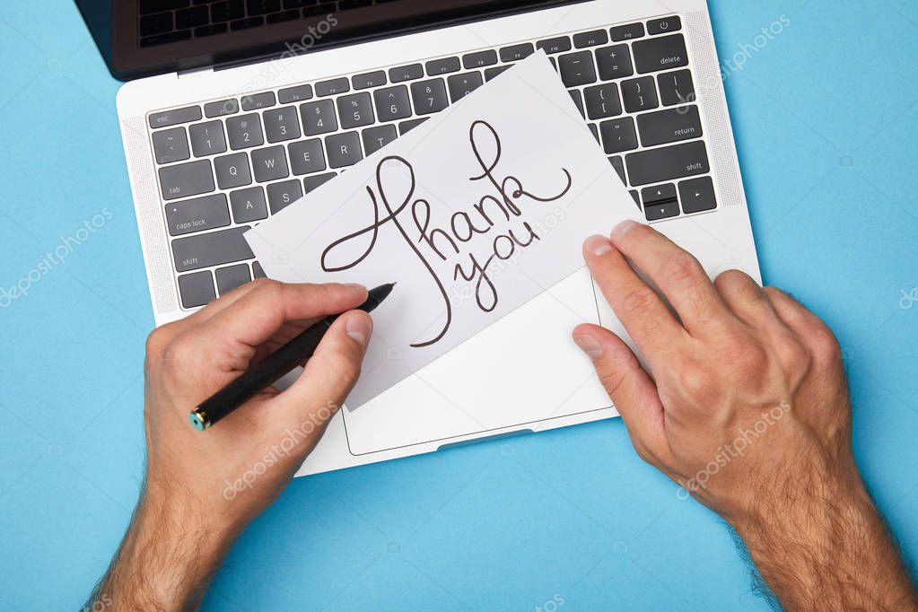 cropped view of man writing on card with thank you lettering near laptop on blue background