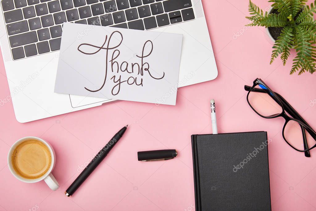 top view of laptop, green plant, coffee, notebook, glasses and card with thank you lettering on pink background