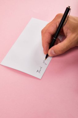 partial view of man writing with pen thank you lettering on white card on pink background clipart