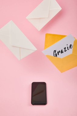 yellow envelope with white card with grazie lettering near letters and smartphone with blank screen on pink background clipart