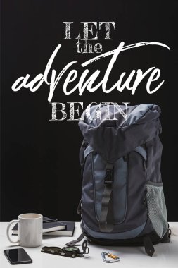 backpack, cup, notebooks, smartphone and trekking equipment isolated on black with let the adventure begin lettering clipart