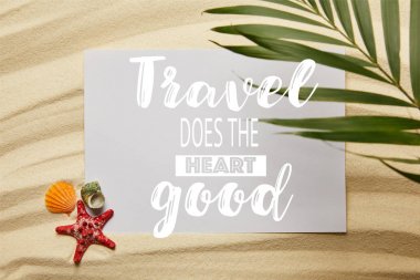 top view of green palm leaf near placard with travel does the heart good, starfish and seashells on sandy beach  clipart