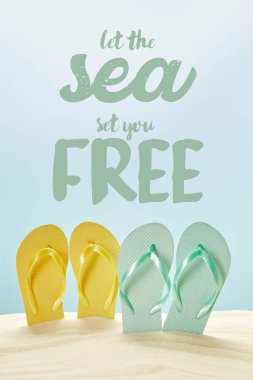 summer yellow and blue flip flops in golden sand isolated on blue with let the sea set you free lettering clipart