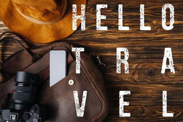 top view of brown leather bag, hat, digital camera and smartphone on wooden table with hello travel illustration