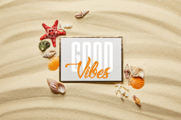 top view of placard with good vibes lettering near seashells, starfish and white corals on sandy beach 