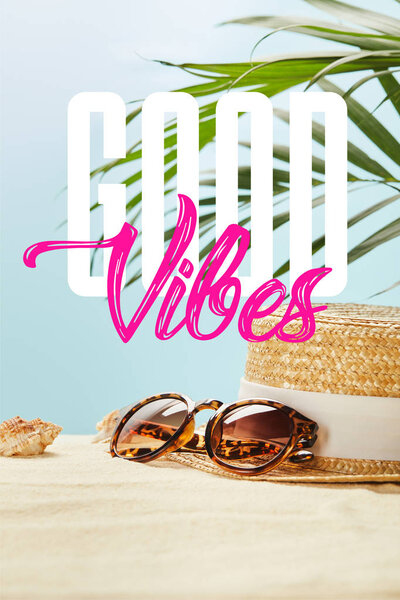 sunglasses near straw hat and seashells in summertime isolated on blue with good vibes lettering