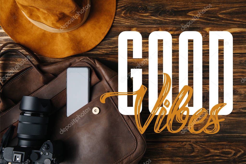 top view of brown leather bag, hat, digital camera and smartphone on wooden table with good vibes illustration