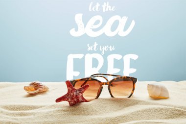brown stylish sunglasses on sand with seashells and starfish on blue background with let the sea set you free lettering clipart