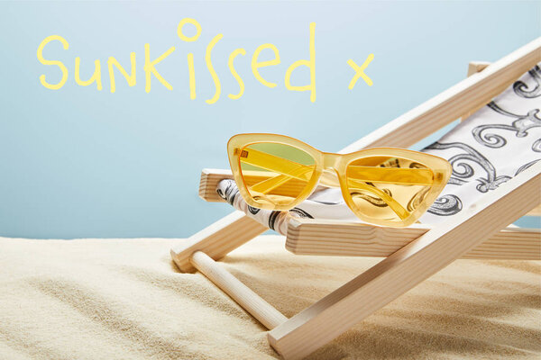 желтые стильные солнцезащитные очки на шезлонге на песке на синем фоне с надписью
