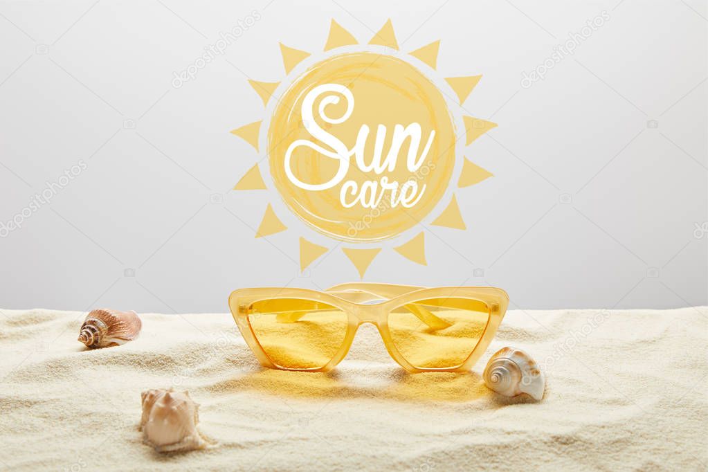 yellow stylish sunglasses on sand with seashell on grey background with sun care lettering