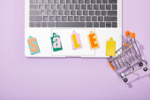 top view of colorful shopping bags with sale lettering on laptop near small toy cart on violet 
