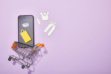 mor üzerinde oyuncak sepetine düşen giysi ve sarı kağıt torba kağıt kesim ile akıllı telefonun üst görünümü 