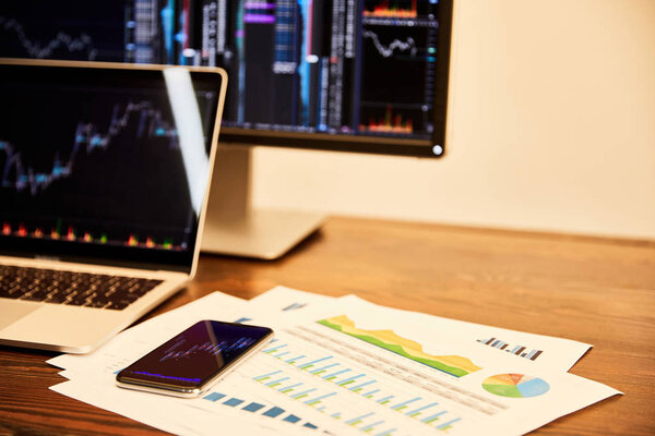 ноутбук и компьютер с диаграммами и графиками, документами и смартфоном на столе
  