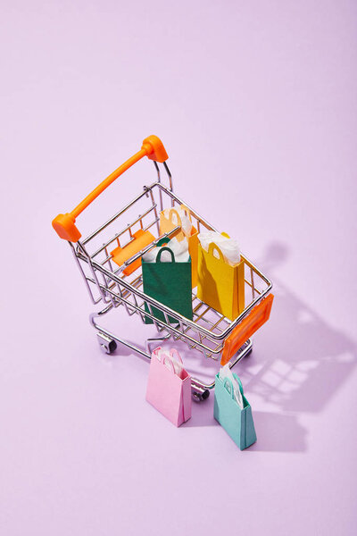few small paper bags near toy cart with colorful shopping bags on violet background