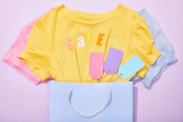 top view of sale lettering on t-shirts with colorful tags in shopping bag on violet