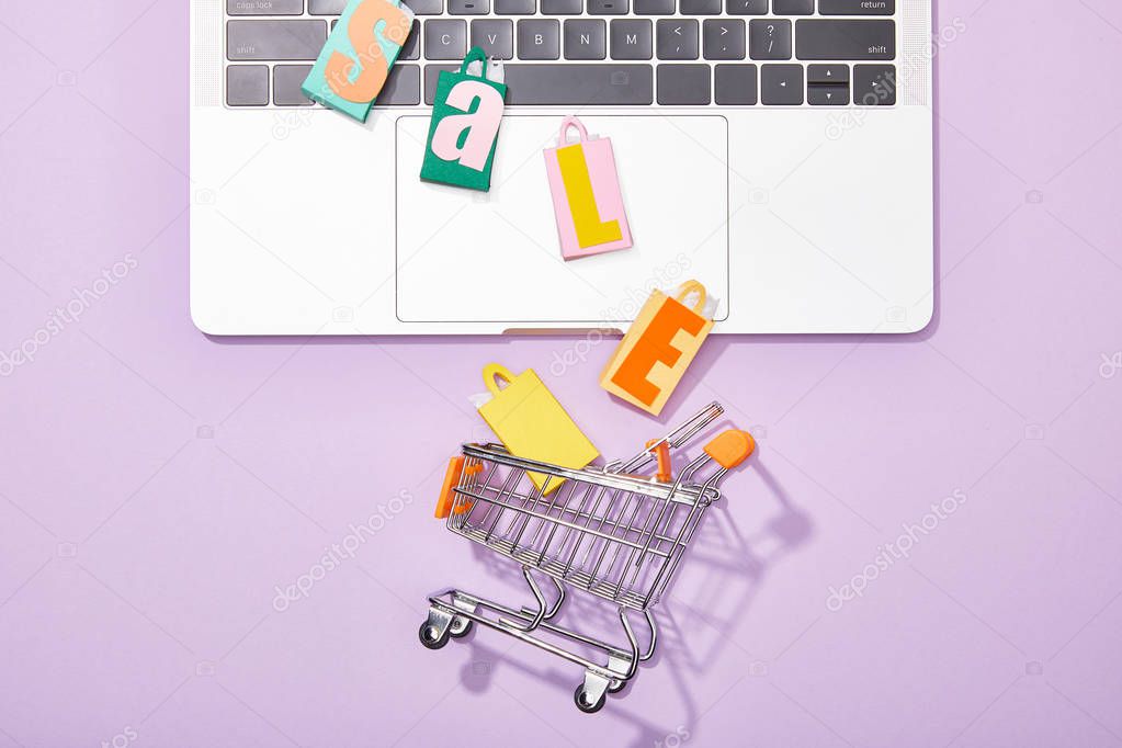top view of colorful toy bags with sale lettering on laptop falling in toy shopping cart on violet 