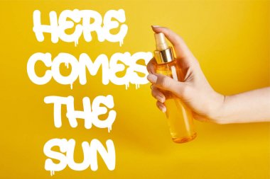 cropped view of woman holding bottle with sunscreen spray on yellow background with here comes the sun lettering clipart