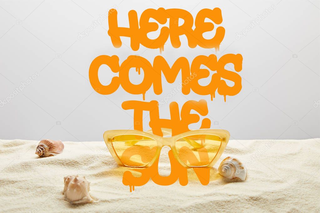yellow stylish sunglasses on sand with seashell on grey background with here comes the sun lettering